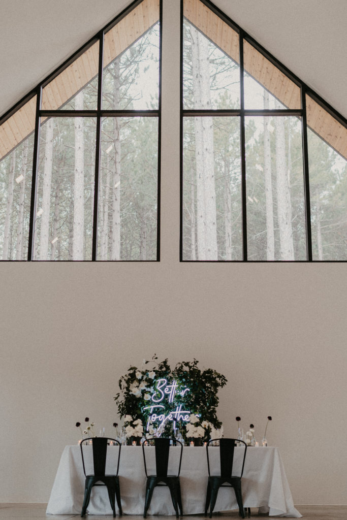 An modern sweetheart table with neon sign and greenery backdrop.